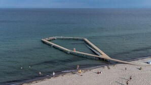 Ein Strand mit einer markanten, geometrischen Pier, die sich ins Meer erstreckt, umgeben von einem sandigen Ufer, an dem sich Menschen aufhalten.