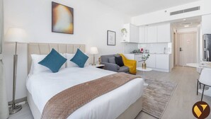 Een moderne hotelkamer met een groot bed, een grijze bank, een klein rond tafeltje en een kitchenette op de achtergrond.