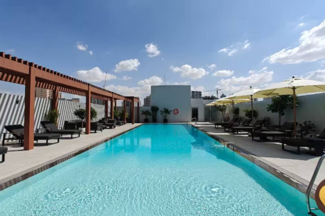 Piscina con sillas de descanso y sombrillas, rodeada por una pérgola y un edificio blanco.