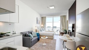 A modern hotel room with a kitchenette, a sofa, a bed, a dining area, and a balcony with a view.
