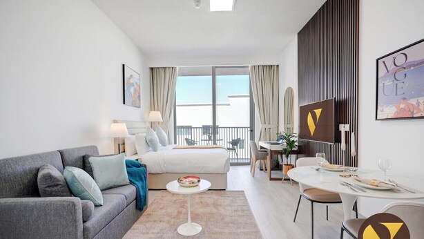 A modern hotel room with a grey sofa, a bed with white bedding, a dining area with a table and chairs, and a balcony with a view.