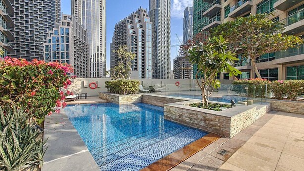 Una piscina en la azotea con un diseño azul turquesa, rodeada de edificios modernos y abundante vegetación.