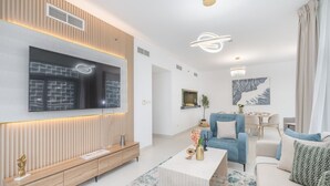 Una sala de estar moderna con un televisor de pantalla plana grande, un mueble bajo de madera, un sillón azul, un sofá blanco y una araña.