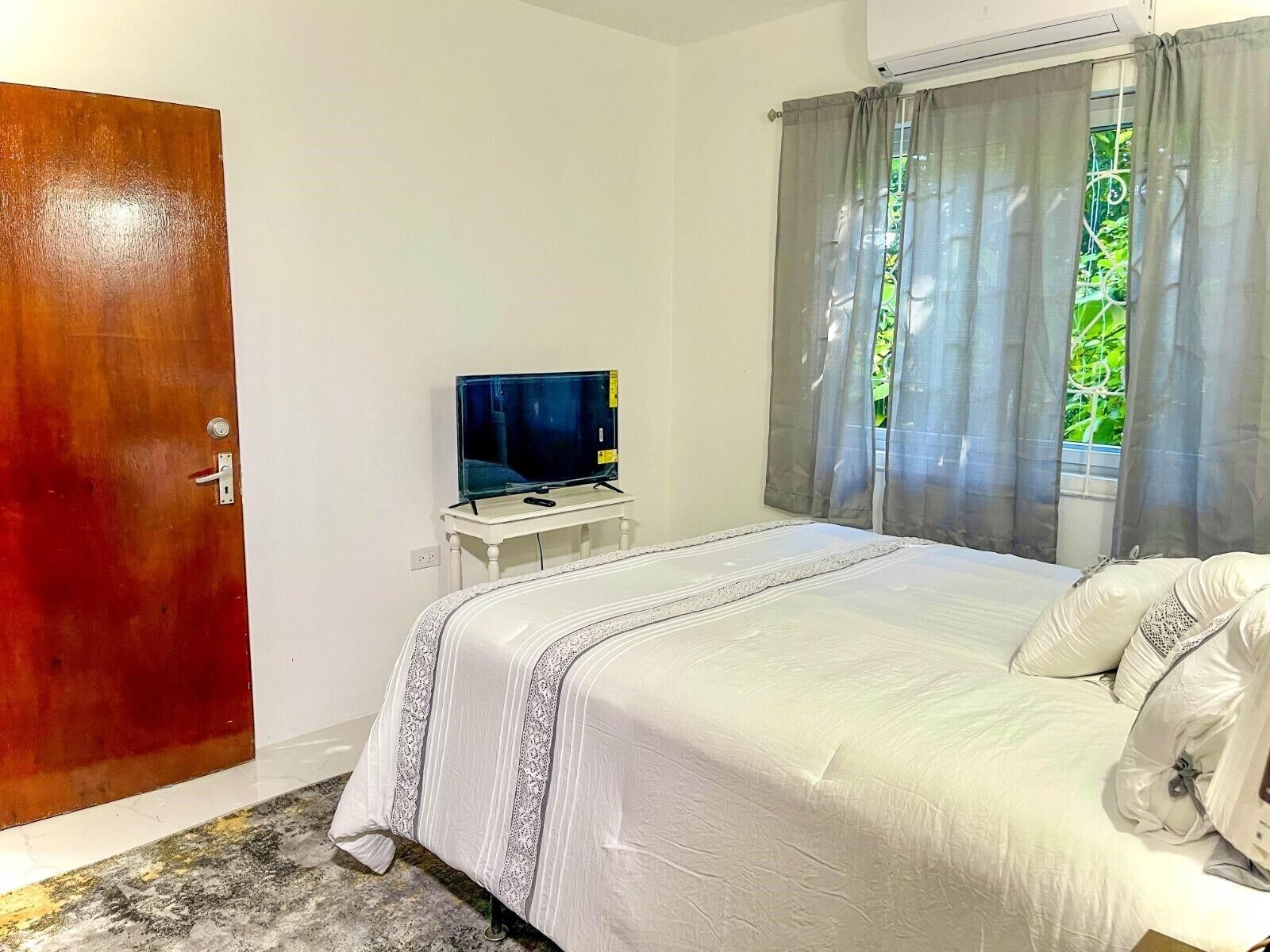 A bedroom with a bed, a television on a small table, a wooden door, and a window with curtains.
