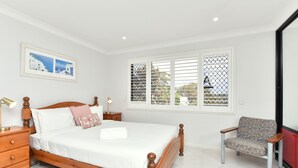 A neatly arranged bedroom with a wooden bed, a bedside table with a lamp, a chair, and a window with blinds. - Coastal Bliss Studio at Shoal Bay (Shoal Bay)