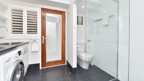 A modern bathroom with a glass shower enclosure, a Bosch washing machine, and a wooden door with blinds. - Coastal Bliss Studio at Shoal Bay (Shoal Bay)