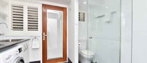 A modern bathroom with a glass shower enclosure, a Bosch washing machine, and a wooden door with blinds.