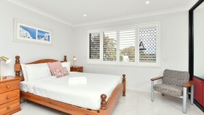 A bedroom with a wooden bed, white bedding, a nightstand with a lamp, a chair, and a window with blinds.