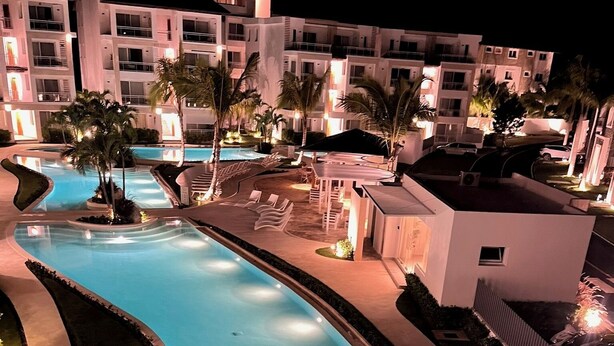 Vista nocturna de un área de piscina de hotel con piscina curvo, sillas de descanso y palmeras.