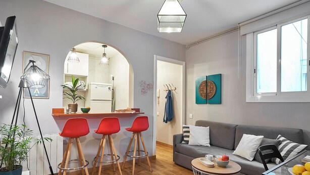 Een moderne woonkamer met een grijze bank, rode barstoelen, een ronde salontafel en een keukenhoek met een koelkast en een hanglamp.