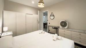 A hotel room with a large bed, a bedside table with a lamp, a dresser, a fan, and a wardrobe.
