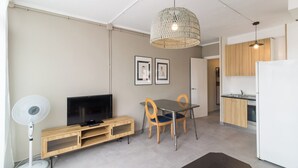 A modern living space with a flat-screen TV, wooden entertainment unit, dining table with chairs, a hanging woven light fixture, and a kitchen area with wooden cabinets and a white refrigerator. - Valles Atico · One bedroom apartment with terrace (Barcelona)