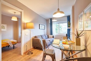 A cozy living room with a sofa, a glass dining table with chairs, a floor lamp, and a window with curtains.
