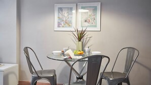 A dining area with a glass table, metal chairs, a vase with plants, and two framed artworks on the wall.