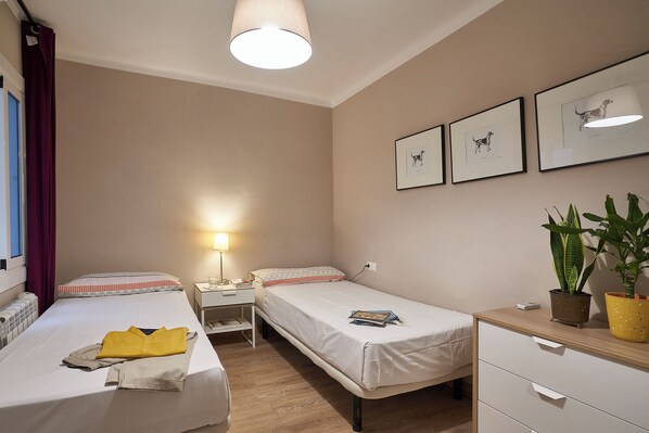 A hotel room with two beds, a bedside table with a lamp, a small desk, and a potted plant.