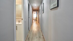 A hallway with a wooden floor, light gray walls, and a ceiling light. There are framed pictures on the wall and a door leading to a bathroom.