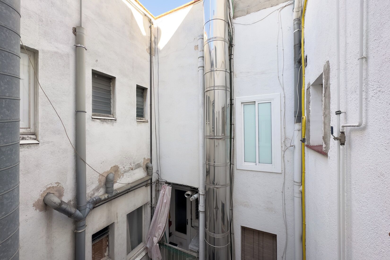 Un vialetto stretto fiancheggiato da edifici, con tubature a vista e una finestra con vetro satinato.