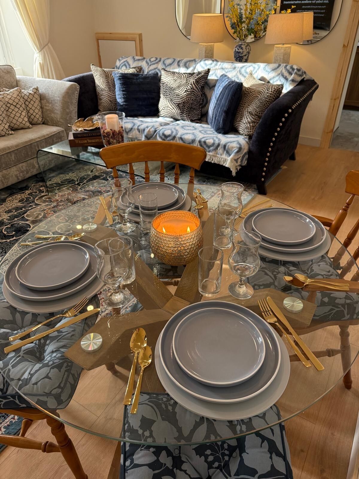 Mesa de comedor preparada para seis personas con superficie de vidrio, rodeada por un tapete estampado, con un sofá y lámparas al fondo.