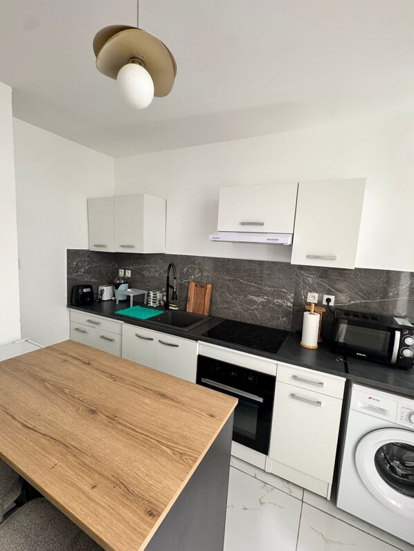 Une cuisine moderne avec des meubles blancs, un plan de travail noir, une table à manger en bois, un four encastré, un four à micro-ondes et un lave-linge.