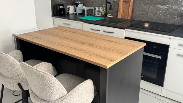 A compact kitchen with a wooden table, two chairs, a black sink, a stove, and white cabinets. - Welcome to MBA HOUSE - Relaxation & Comfort in Guadeloupe! (Pointe-à-Pitre)