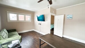 A living room with a green couch, a glass coffee table, a wooden dining table, a ceiling fan, and a TV mounted on the wall. - Downtown Historical 1 Bed Apt (Bay City)