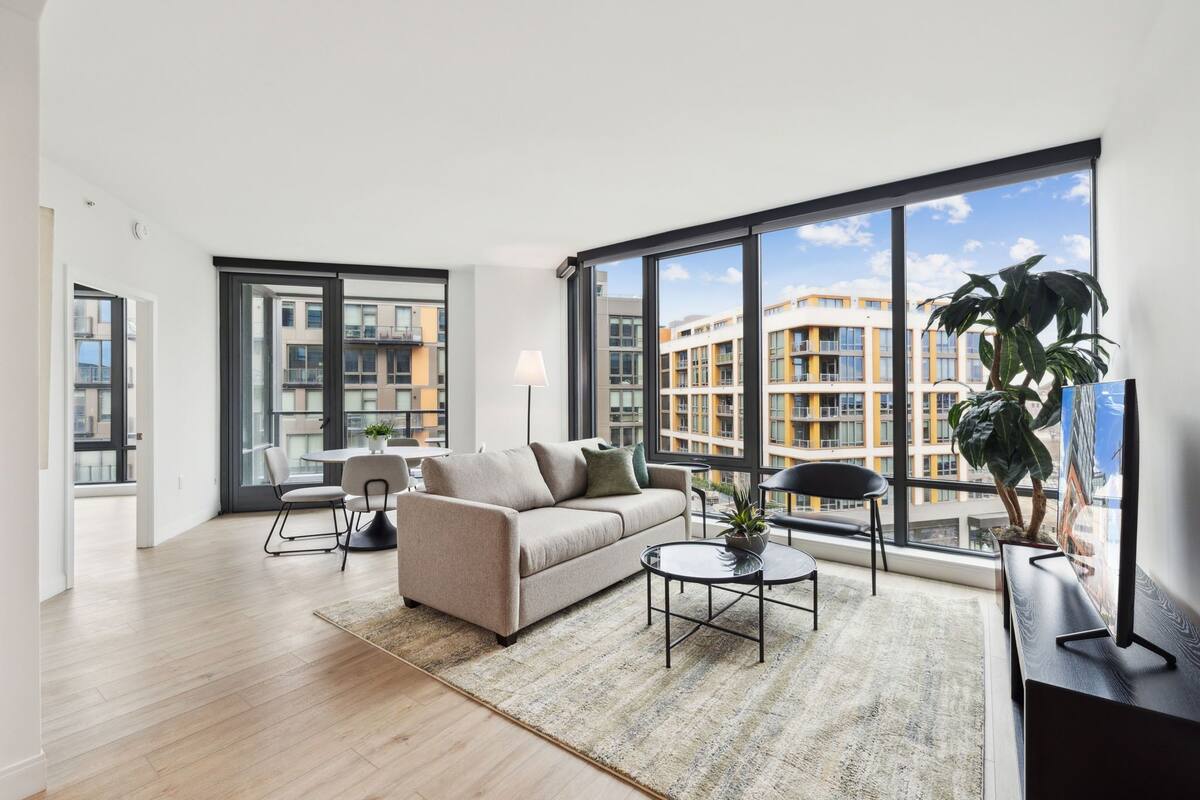 A modern living room with a sofa, coffee table, and chairs, large windows with a city view, and a potted plant.