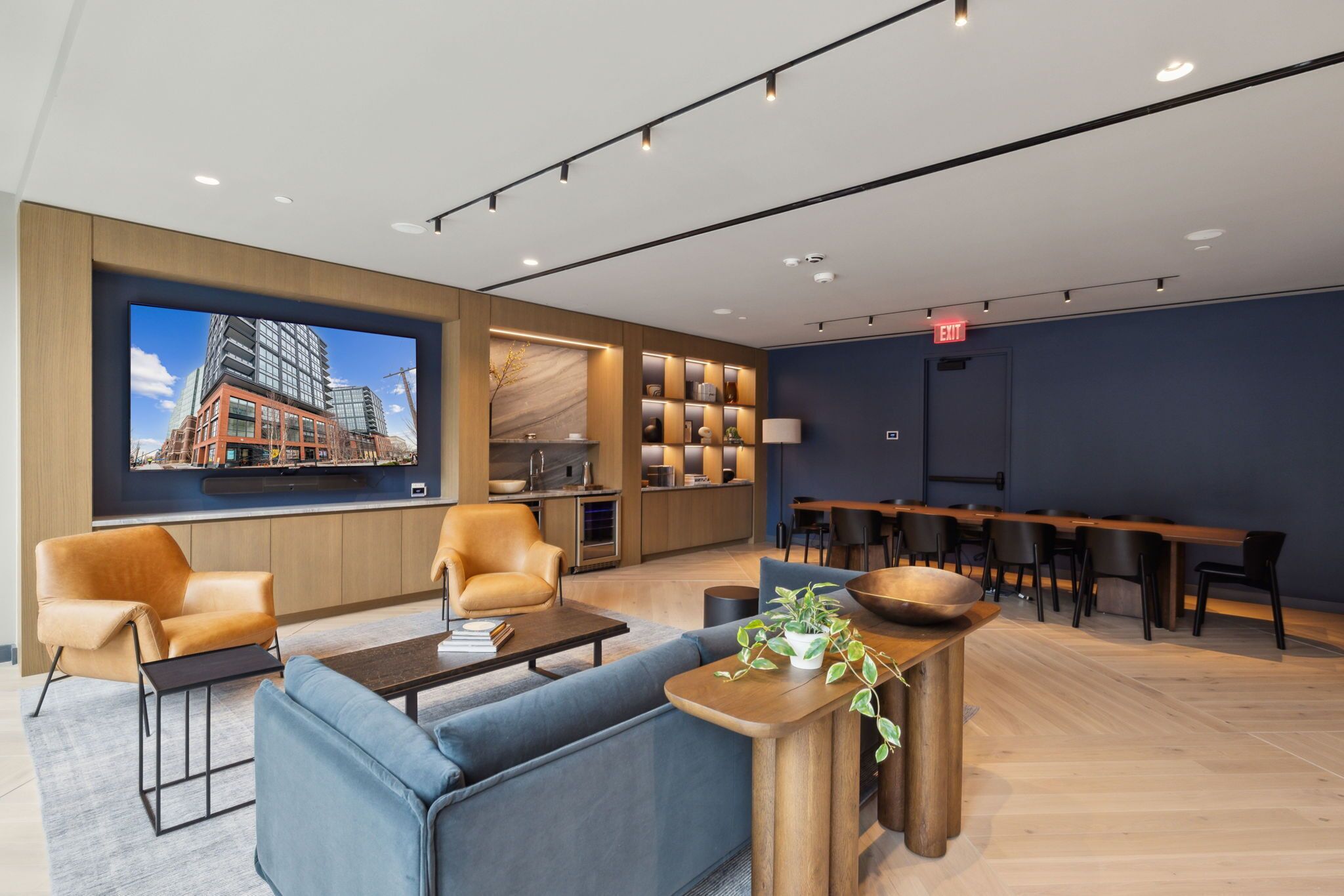 A modern hotel lobby with a large TV screen, comfortable seating, a kitchen area, and a bar.