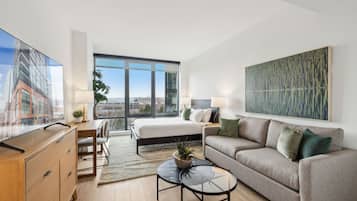 A modern hotel room with a large bed, a sofa, a desk, and a TV. There is a large window with a view of buildings and a painting on the wall.