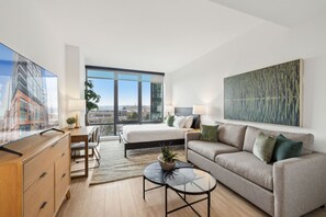 A modern hotel room with a large bed, a sofa, a desk, and a TV. There is a large window with a view of buildings and a painting on the wall. - Placemakr Buzzard Point (Washington)