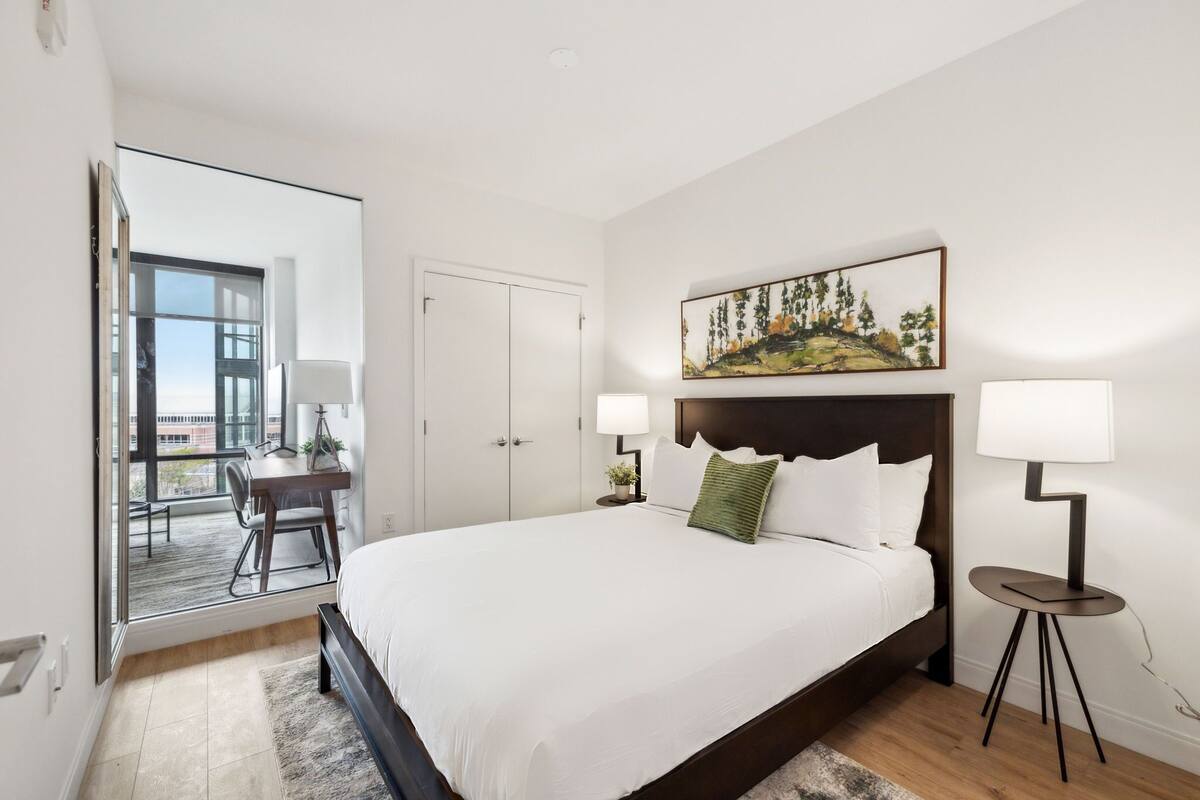 A modern bedroom with a large bed, a desk, and a view through a glass door.