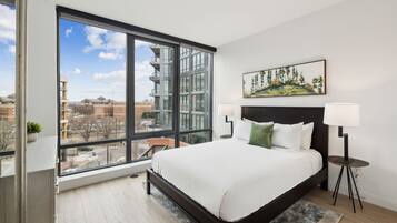 A modern bedroom with a large window, a bed with white bedding, a nightstand, a lamp, and a painting on the wall.