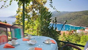 A table set for two with white cups and saucers, orange napkins, and a view of a coastal landscape with buildings and boats.