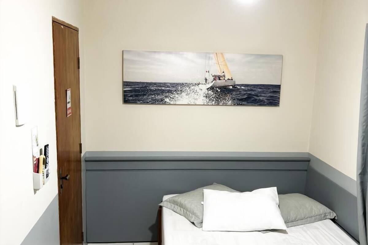 A hotel room with a bed, a ceiling fan, a painting of a sailboat, and a door.
