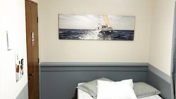 A hotel room with a bed, a ceiling fan, a painting of a sailboat, and a door.