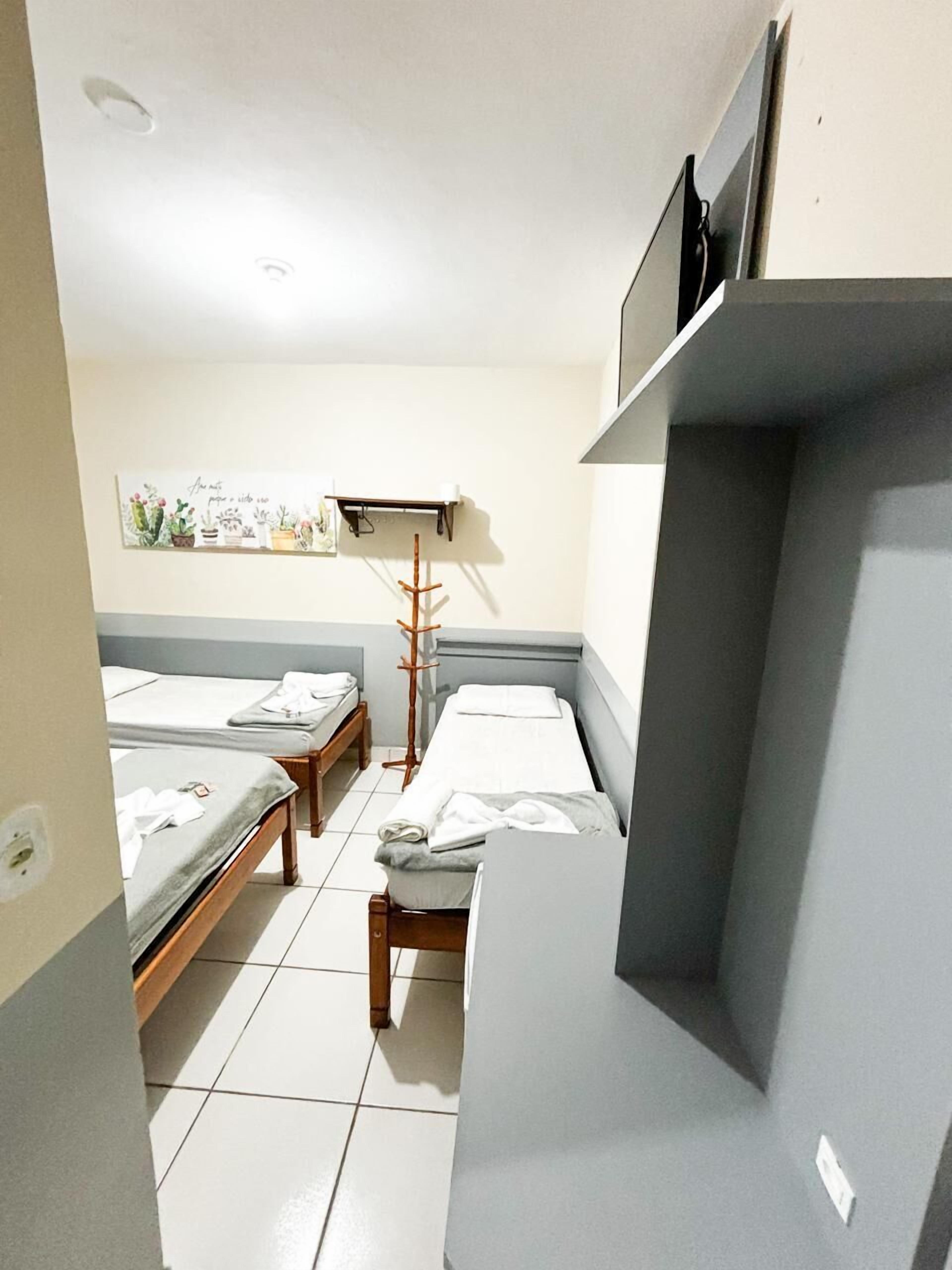 Habitación de hotel compacta con dos camas individuales, una mesita de noche pequeña, una silla, una lámpara de pie y un televisor montado en la pared.