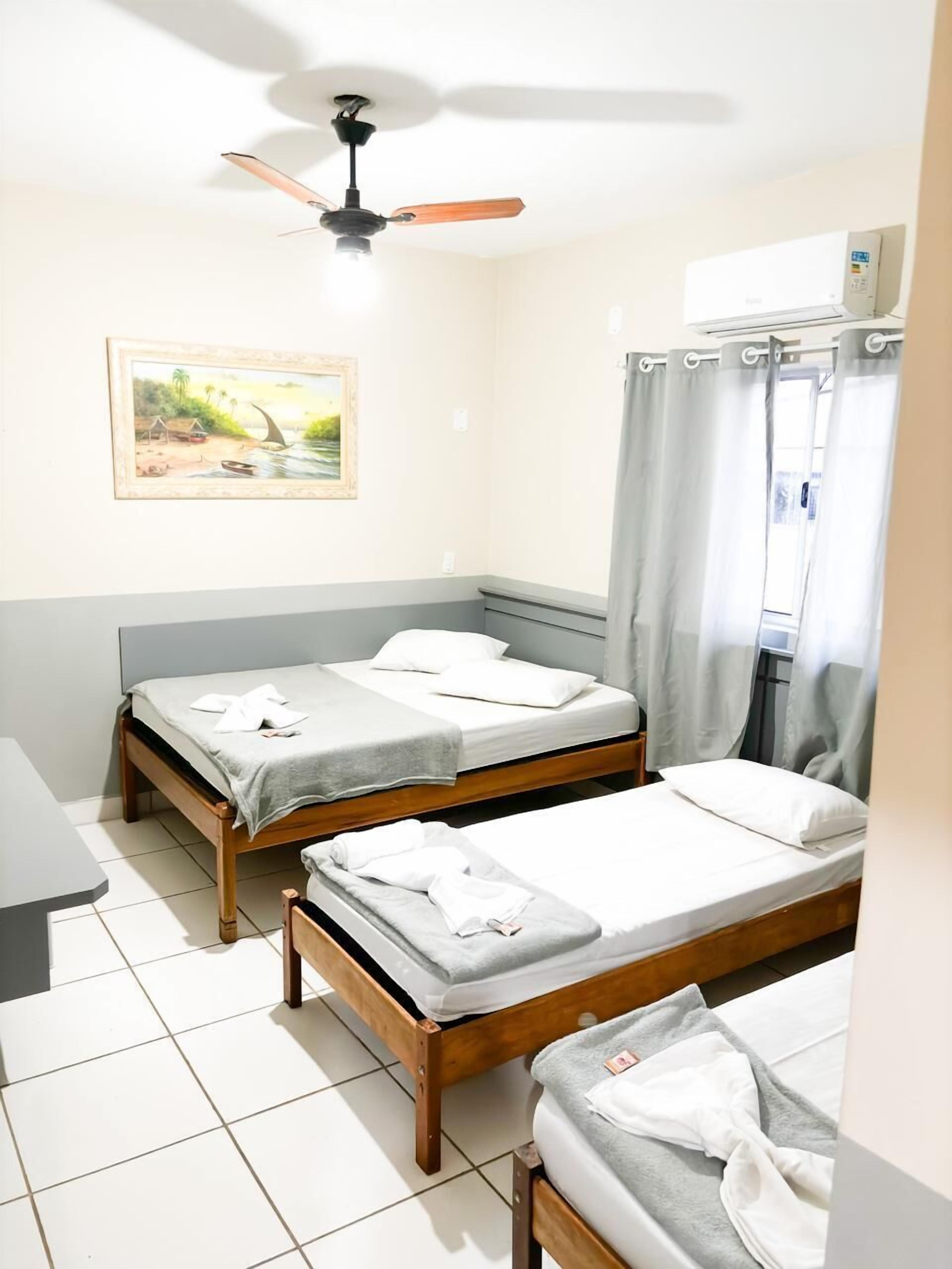 Quarto com duas camas de madeira, um ventilador de teto, janela com cortinas e um quadro na parede.