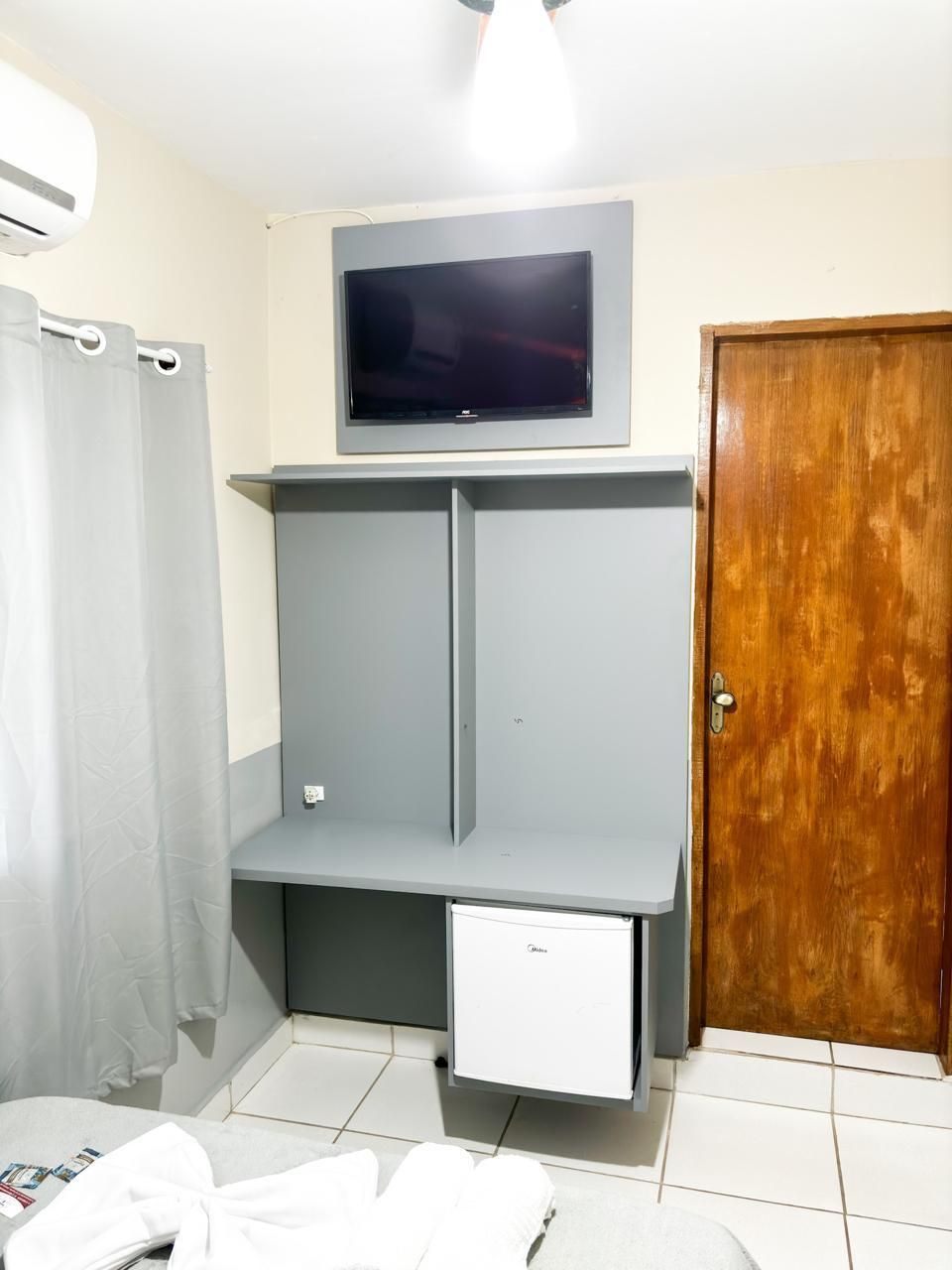 Une pièce de taille modeste, avec une armoire grise, un petit meuble blanc, un téléviseur fixé au-dessus de l’armoire, une porte en bois et un rideau de douche.
