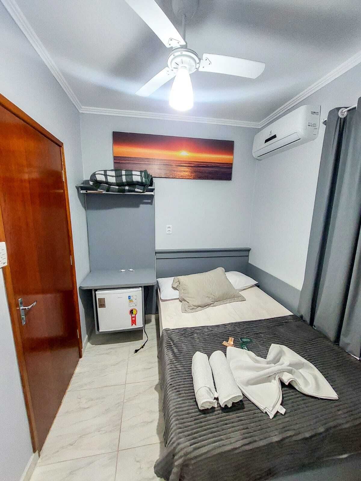 A compact hotel room with a bed, a ceiling fan, an air conditioning unit, a small desk with a computer, and a painting of a sunset.