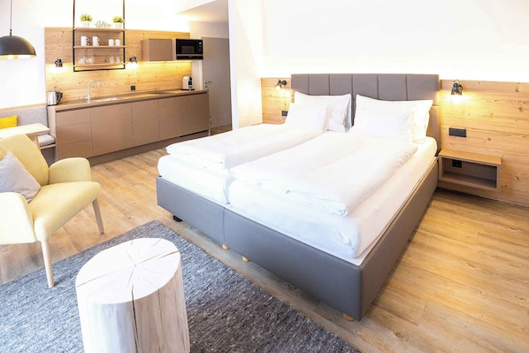 A modern hotel room with a large bed, a small desk, a chair, a wooden nightstand, and a kitchen area with a microwave.
