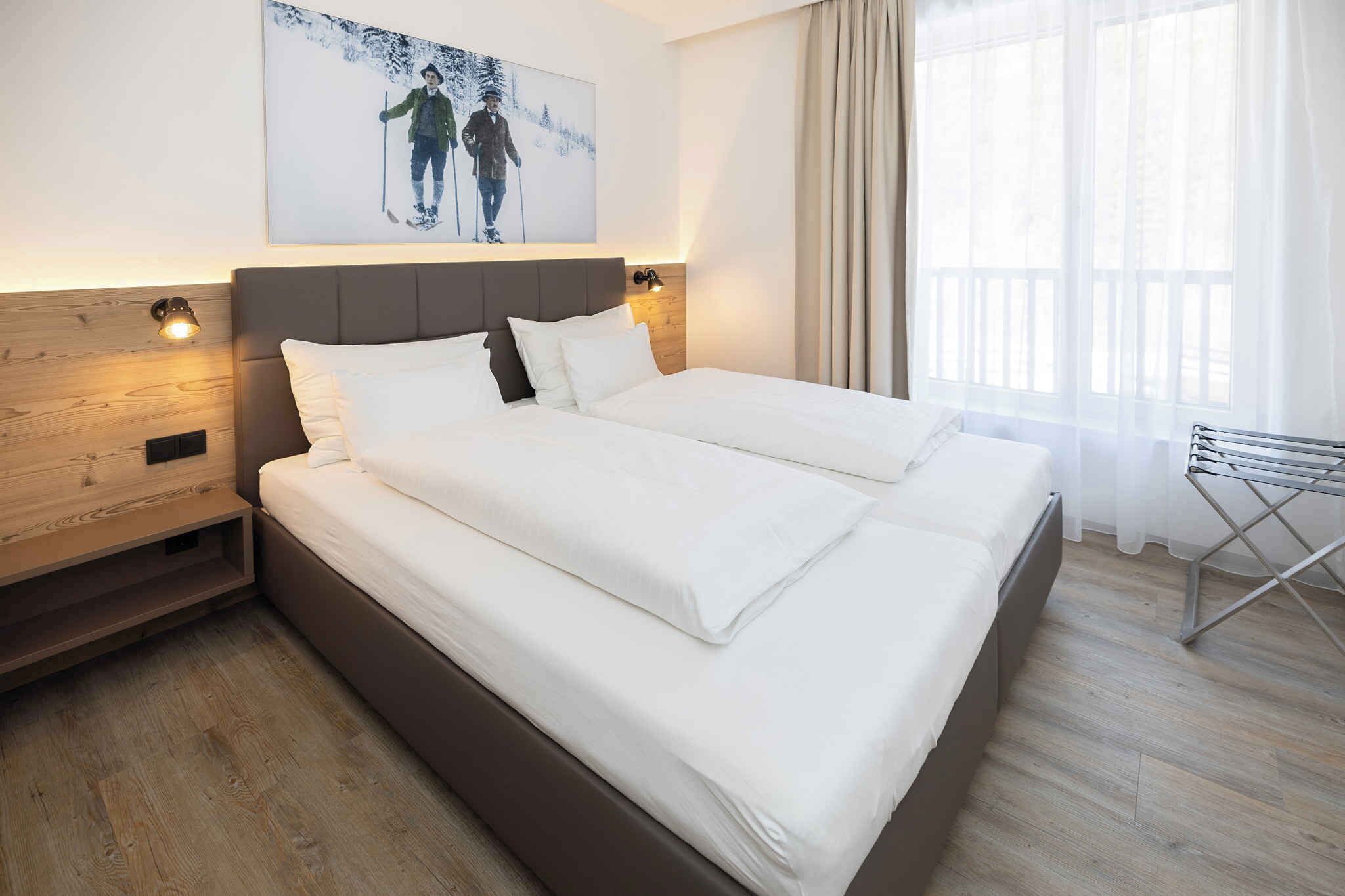 Een hotelkamer met een groot bed, een houten hoofdeinde, twee bedlampen, een schilderij aan de muur en een raam met gordijnen.