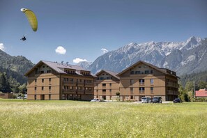 Ein mehrstöckiges Gebäude mit Balkonen, ein Feld im Vordergrund und Berge im Hintergrund.