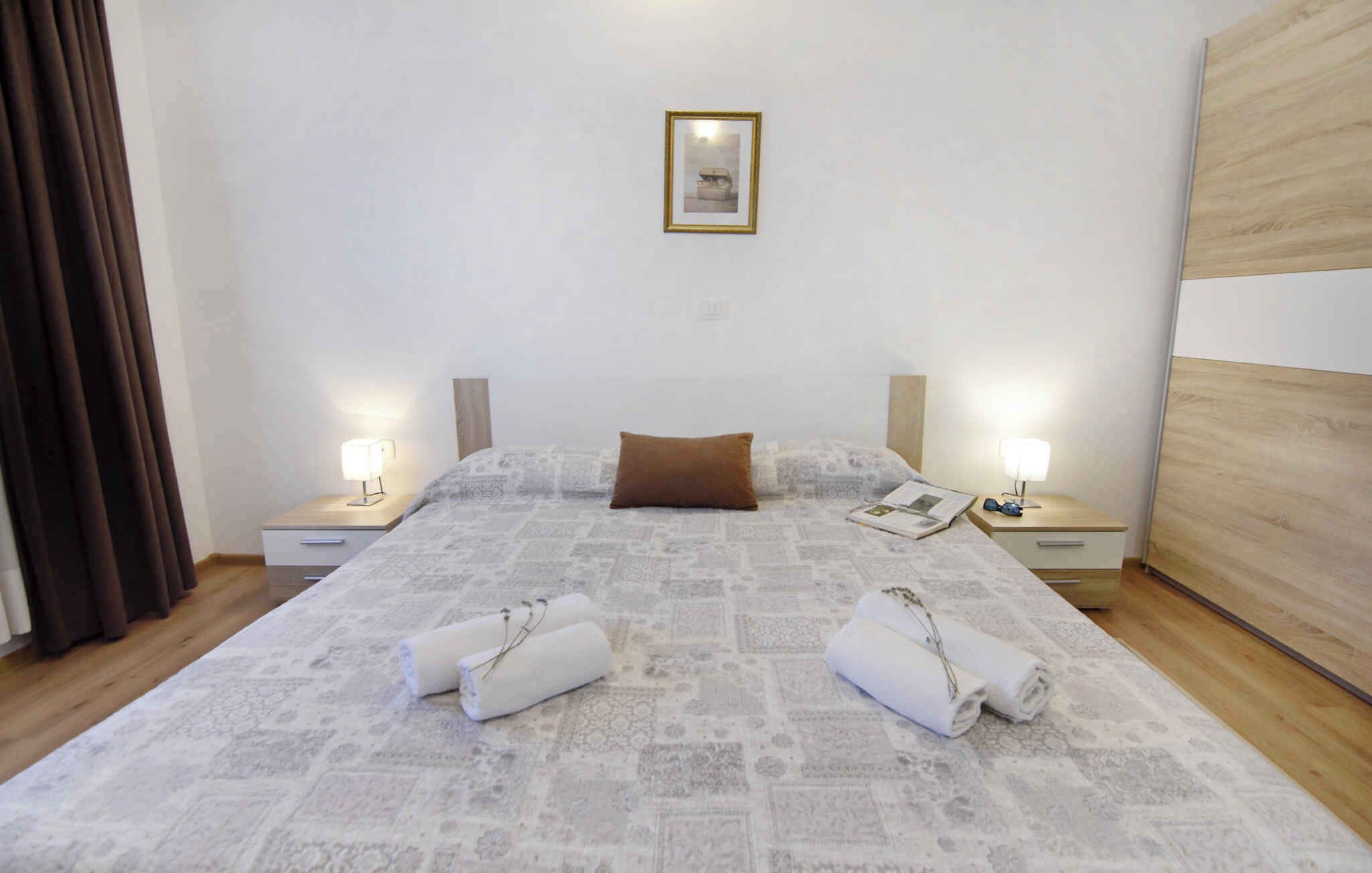 A neatly made bed with a patterned bedspread, two pillows, and two rolled towels on the floor.