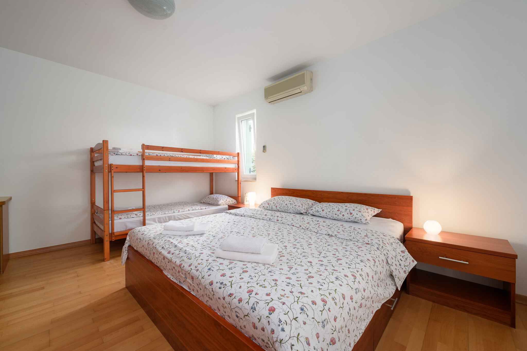 A bedroom with a wooden bunk bed, a single bed with floral bedding, a bedside table with a lamp, and a window with natural light.