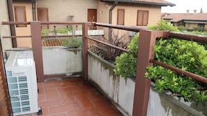 Een smalle buitenpassage met een metalen hekwerk, een betonnen muur met groene planten en een betegelde vloer.