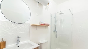 A modern bathroom with a white sink, a toilet, a glass shower enclosure, and a round mirror. - Cosy, modern air-conditioned T2 (Villeurbanne)