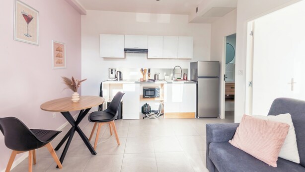 A modern kitchen with white cabinets, a microwave, and a refrigerator. There is a dining area with a wooden table and black chairs. - Cosy, modern air-conditioned T2 (Villeurbanne)