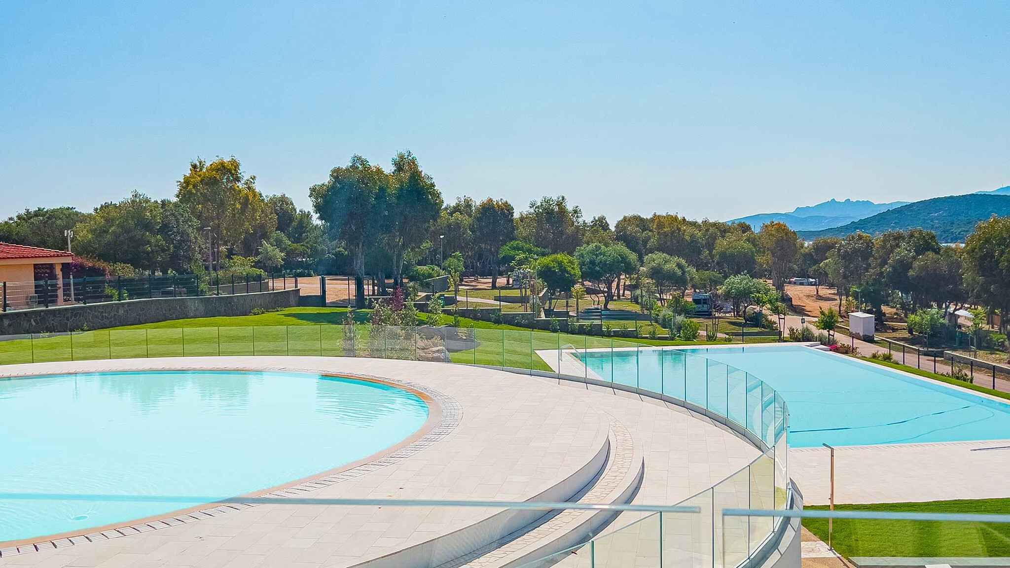 Una piscina de diseño curvo, rodeada por un camino empedrado y un césped con árboles y una cadena montañosa en la distancia.