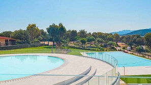A swimming pool with a curved design, surrounded by a paved walkway and a grassy area with trees and a distant mountain range. - Mobile home with pool and near the sea (Le Saline)