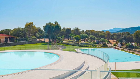 A swimming pool with a curved design, surrounded by a paved walkway and a grassy area with trees and a distant mountain range.