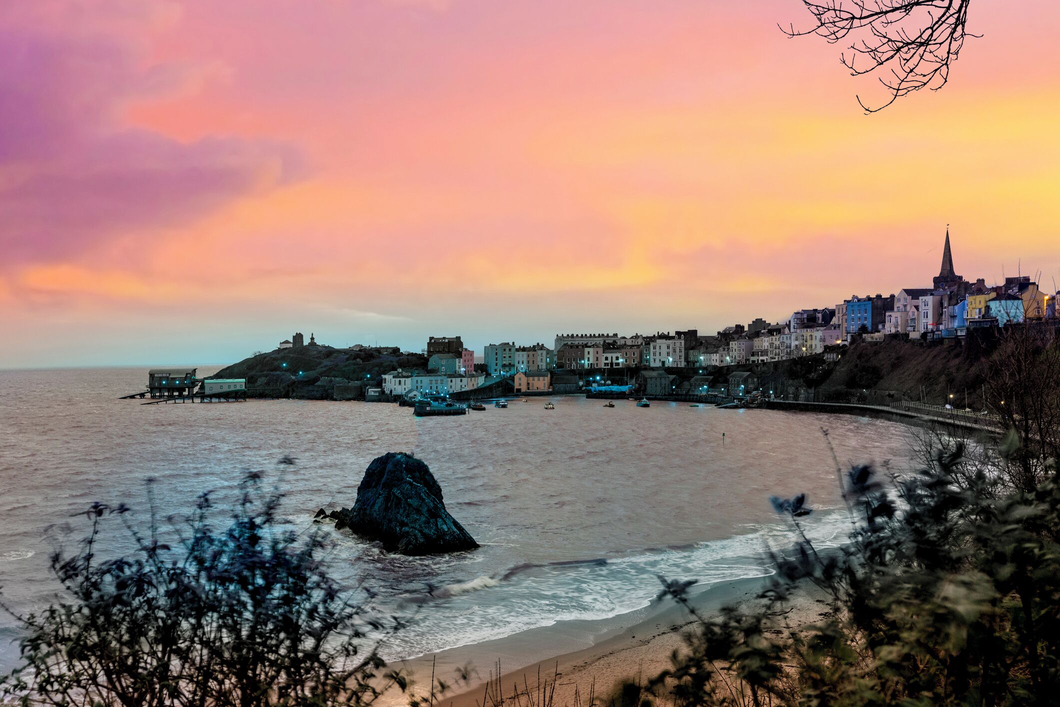 A coastal town with buildings along the shore, a pier, and a prominent rock formation in the water.